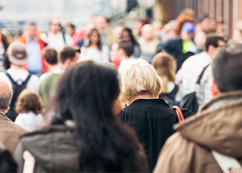 A busy street scene with a diverse crowd of people in various outfits, viewed from behind. The focus is on several individuals walking amongst a blurred background of other pedestrians.